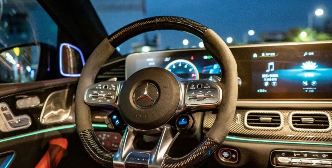 AMG 碳纤维方向盘与碳纤维内饰的成品状态，仪表台上保留了原厂 OLED 旋钮；方向盘总成需与安全气囊与按键功能匹配